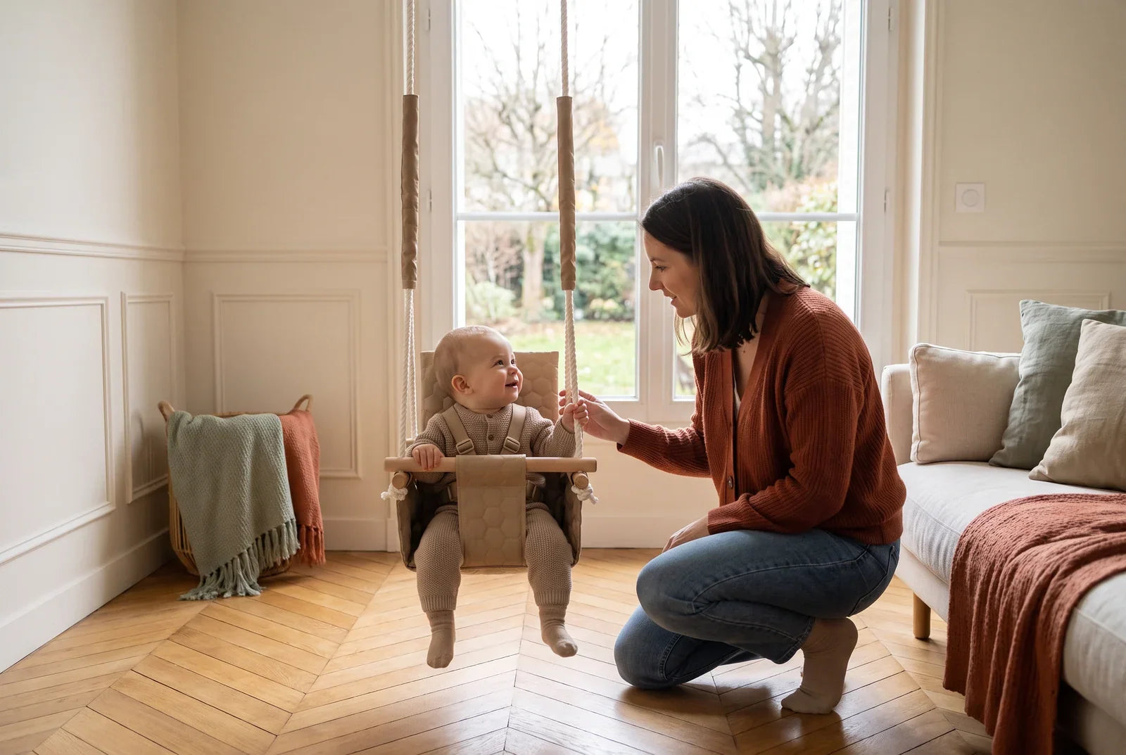 Une maman accroupie près d’un bébé installé dans une balançoire en tissu beige, dans un salon lumineux