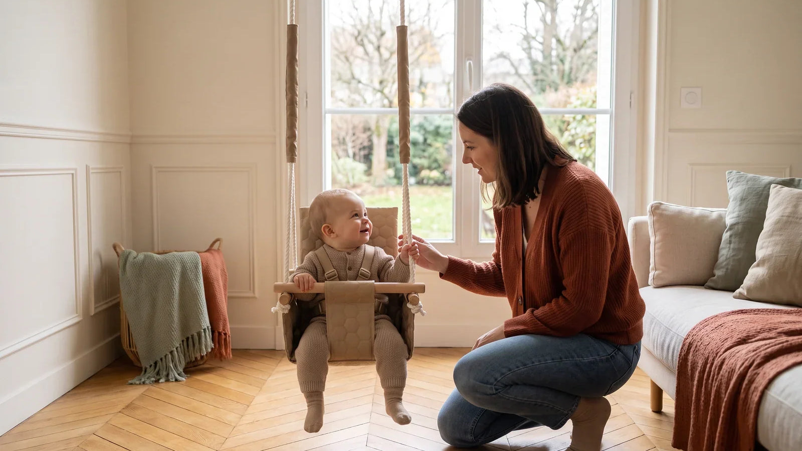 Une maman accroupie près d’un bébé installé dans une balançoire en tissu beige, dans un salon lumineux