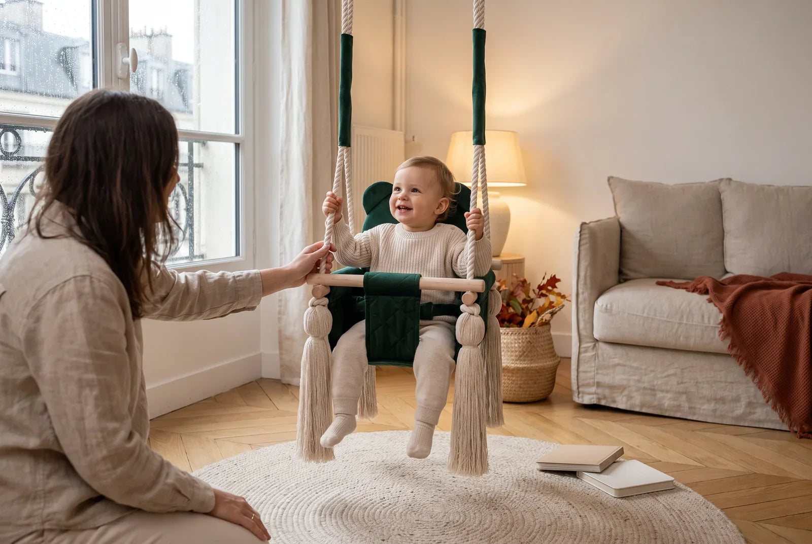 Tout-petit souriant dans une balançoire bébé en velours vert, un adulte à côté, un jour de pluie d’automne