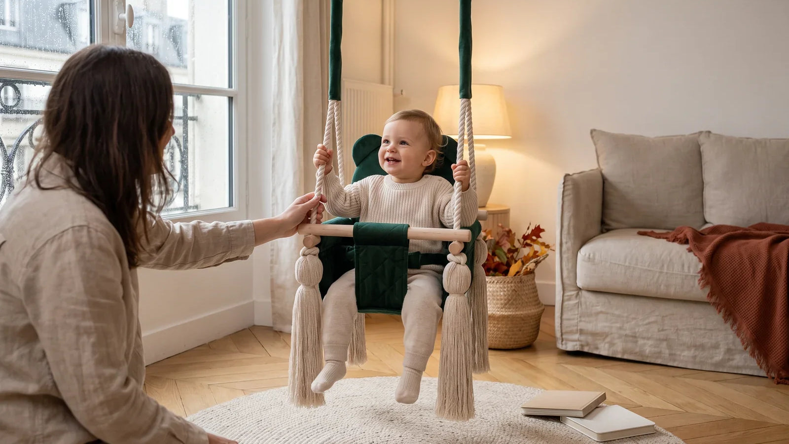 Tout-petit souriant dans une balançoire bébé en velours vert, un adulte à côté, un jour de pluie d’automne