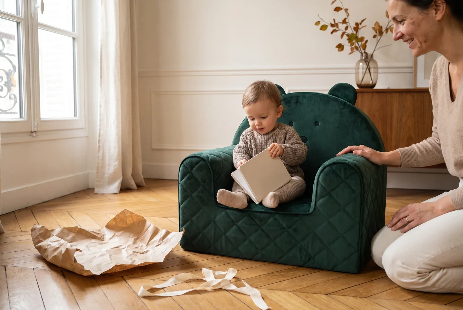 Enfant d’un an assis dans un fauteuil bébé ourson en velours vert avec un livre, un parent agenouillé à côté