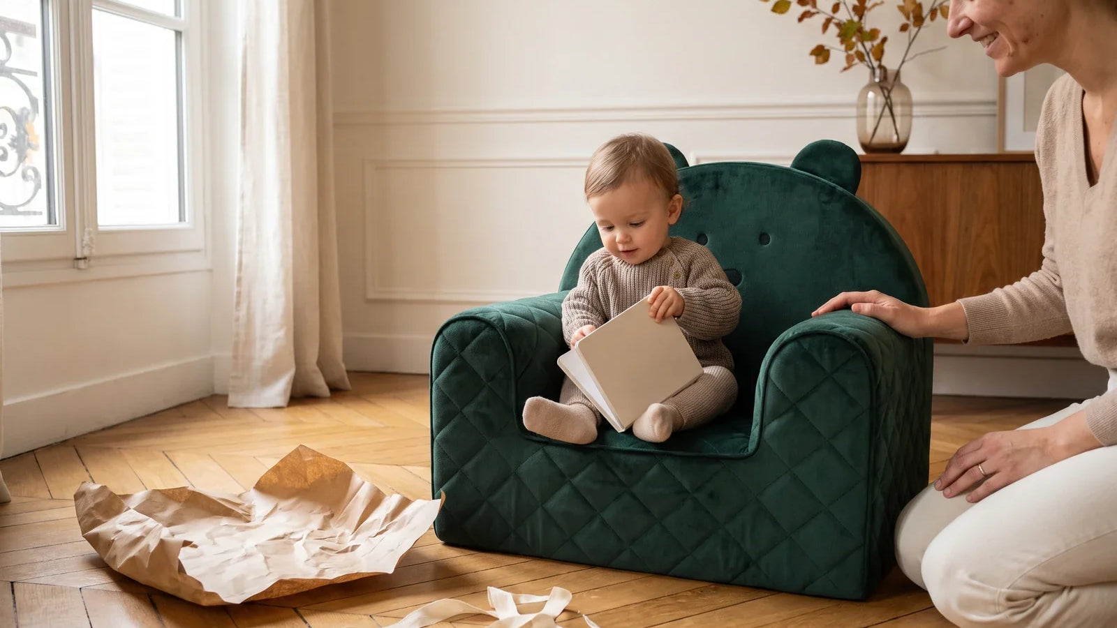 Enfant d’un an assis dans un fauteuil bébé ourson en velours vert avec un livre, un parent agenouillé à côté