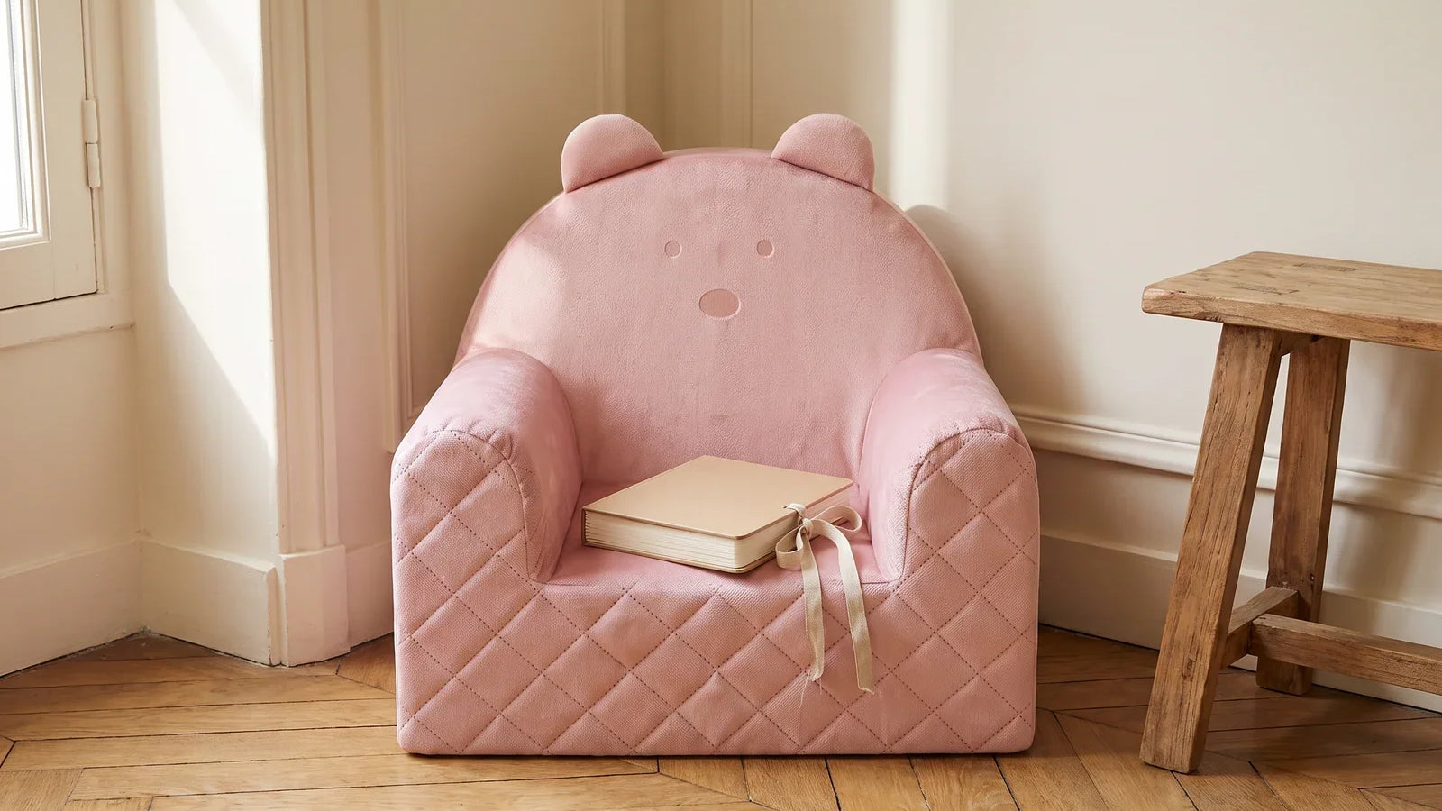 Fauteuil ourson en velours rose dans un coin de chambre lumineux, avec un livre noué d’un ruban posé sur l’assise