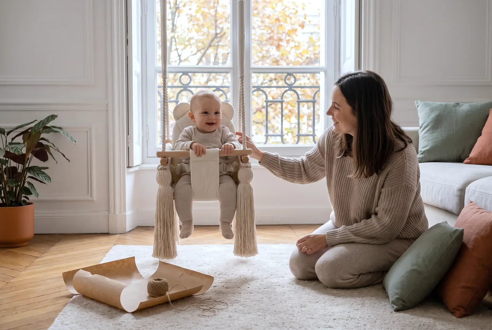 Bébé de 8 mois assis dans une balançoire bébé en velours beige, sa mère à genoux à côté, salon parisien en automne