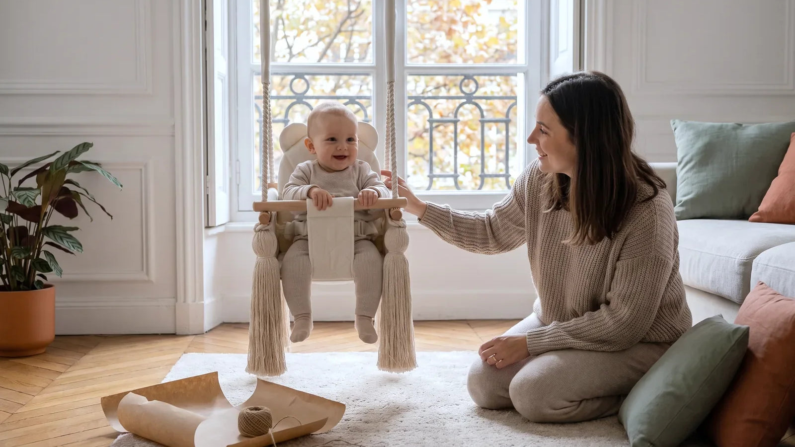 Bébé de 8 mois assis dans une balançoire bébé en velours beige, sa mère à genoux à côté, salon parisien en automne