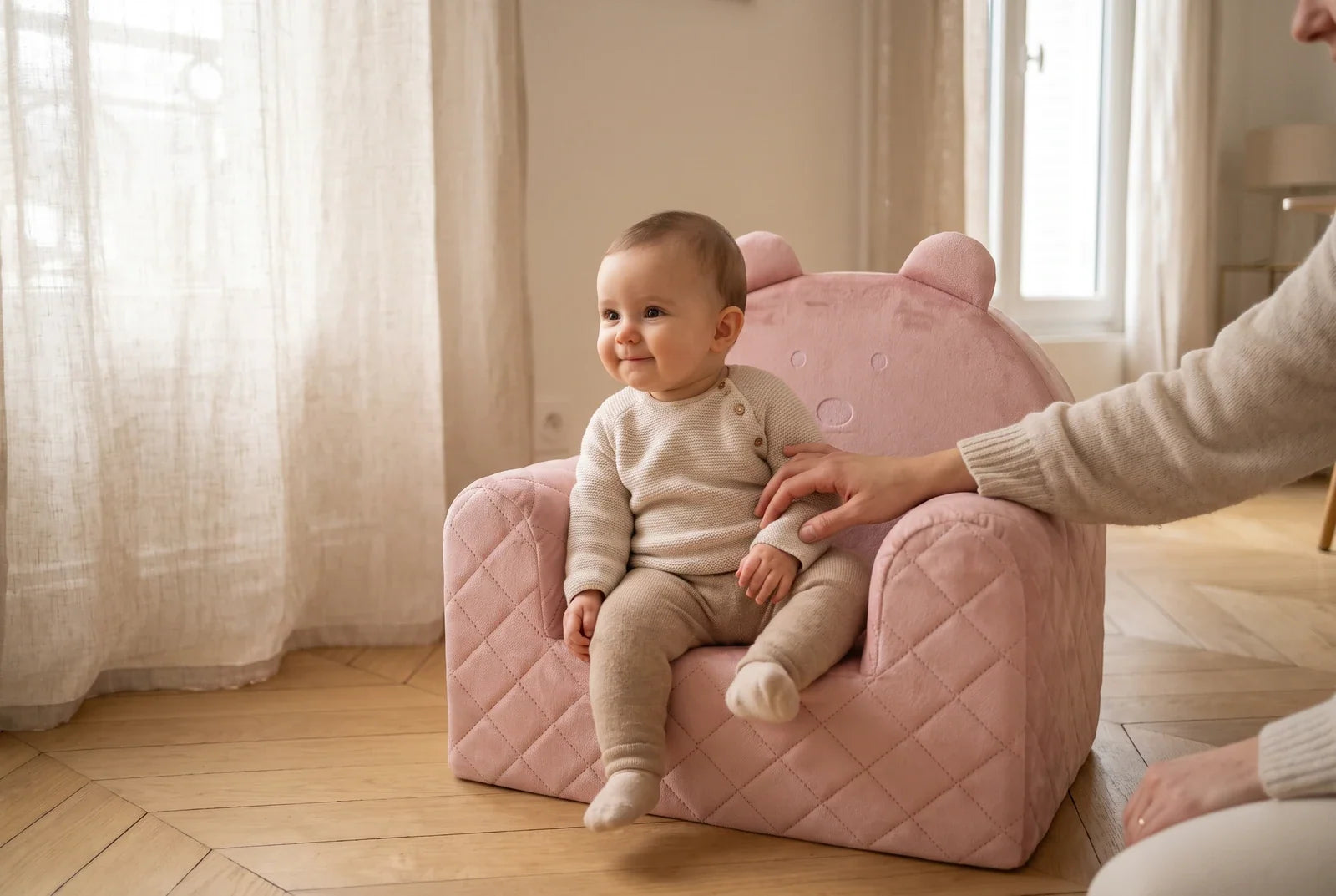 Bébé assis dans un fauteuil ourson en velours rose, un adulte posant la main à côté de lui, dans un salon lumineux