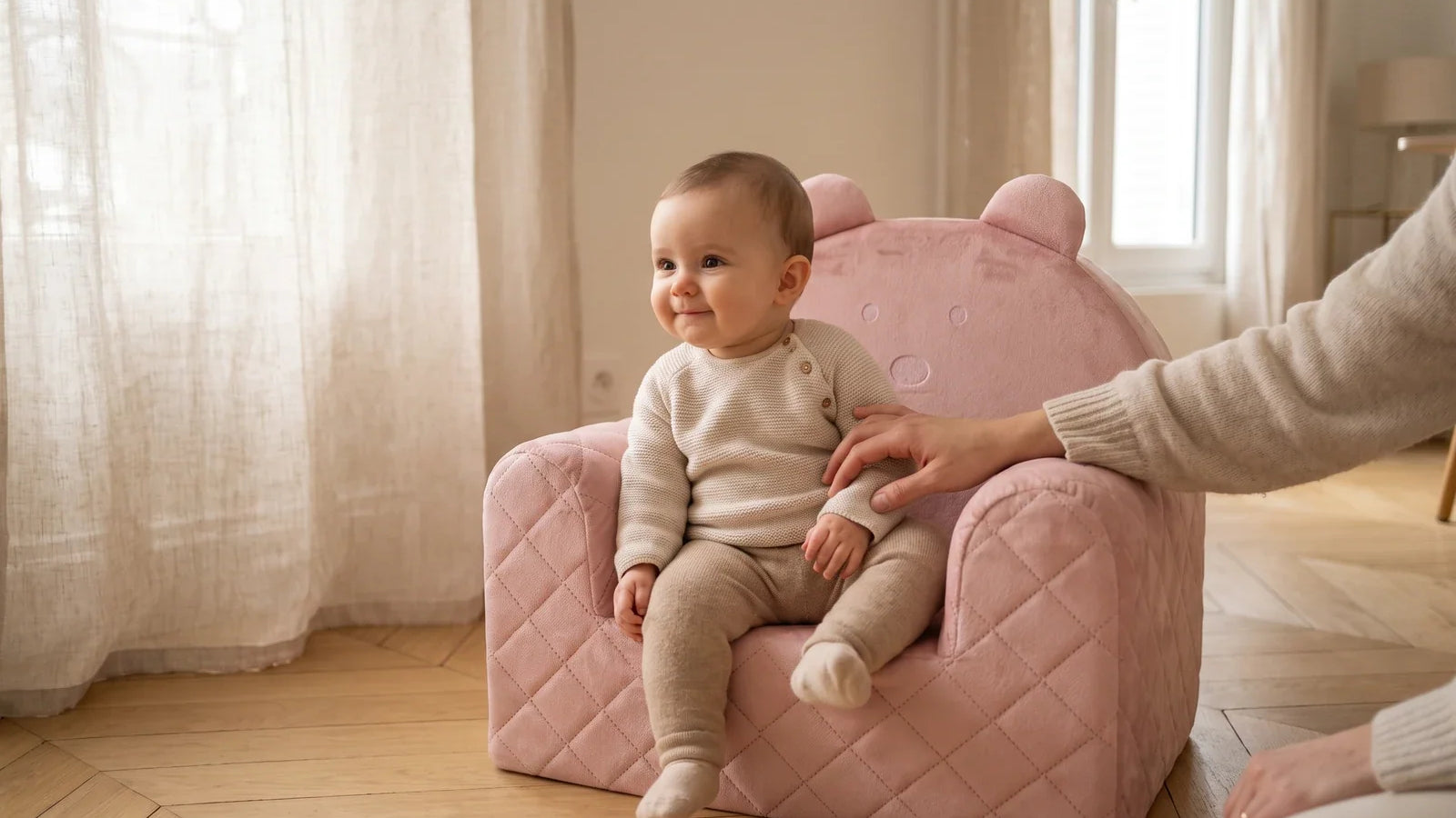 Bébé assis dans un fauteuil ourson en velours rose, un adulte posant la main à côté de lui, dans un salon lumineux