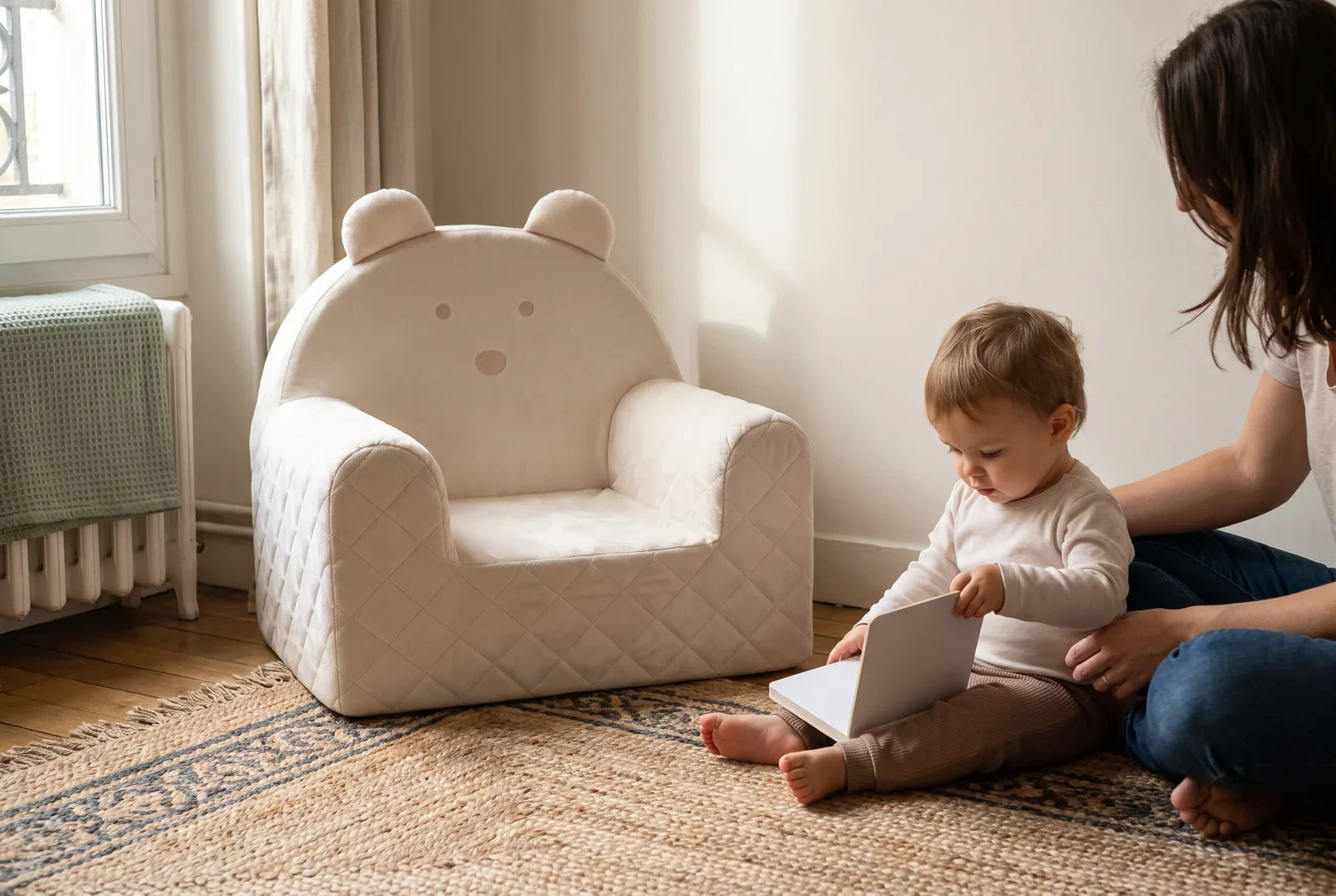 Enfant de 18 mois feuilletant un livre près d’un fauteuil ourson beige, accompagné d’un adulte dans un salon