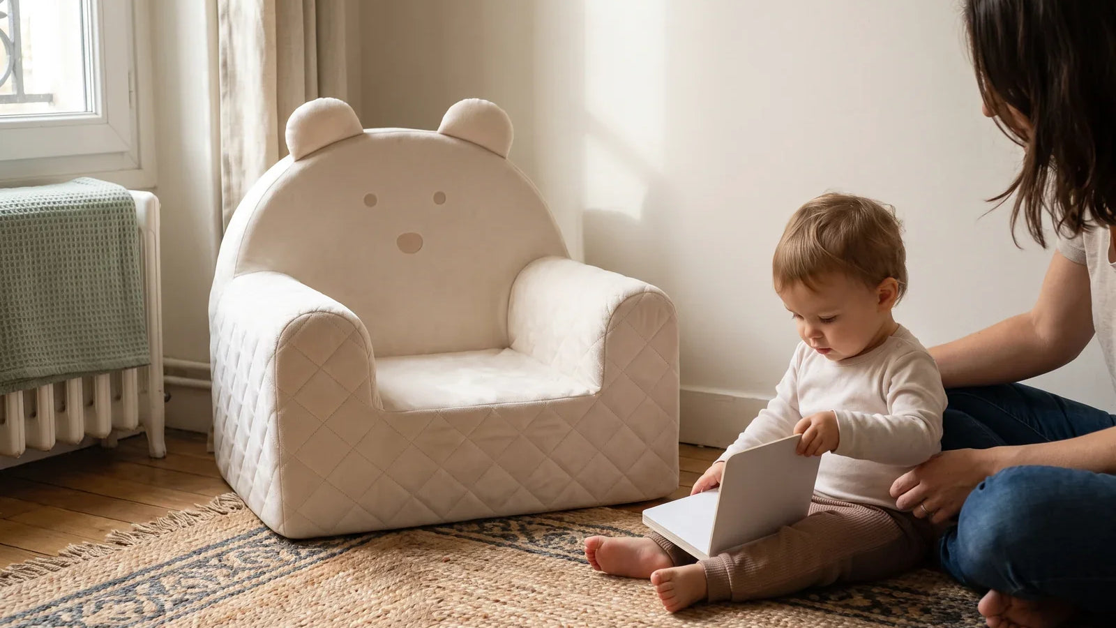 Enfant de 18 mois feuilletant un livre près d’un fauteuil ourson beige, accompagné d’un adulte dans un salon