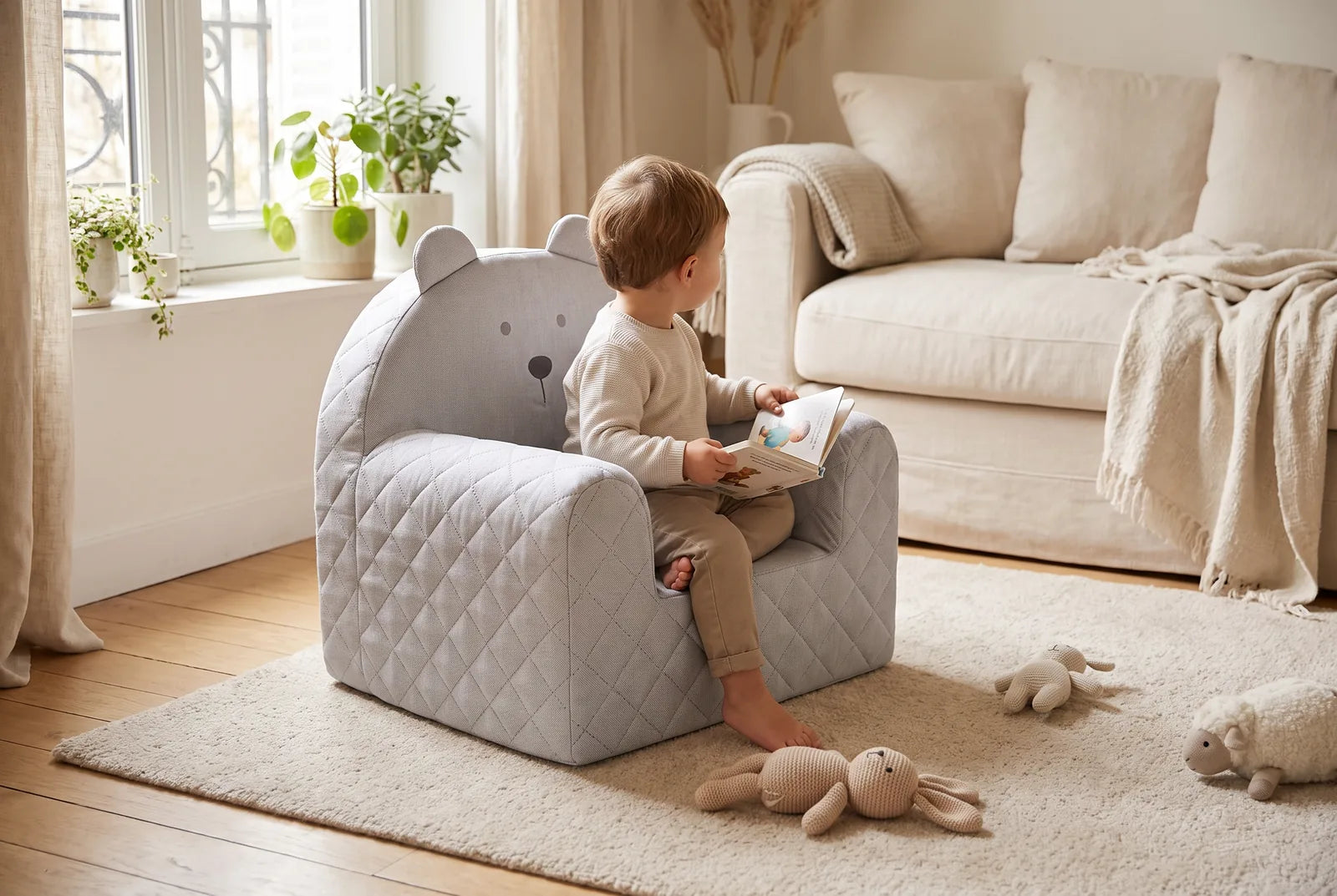 Un enfant de 2 ans et demi lit un livre assis dans un fauteuil gris en forme d’ourson, dans un salon lumineux