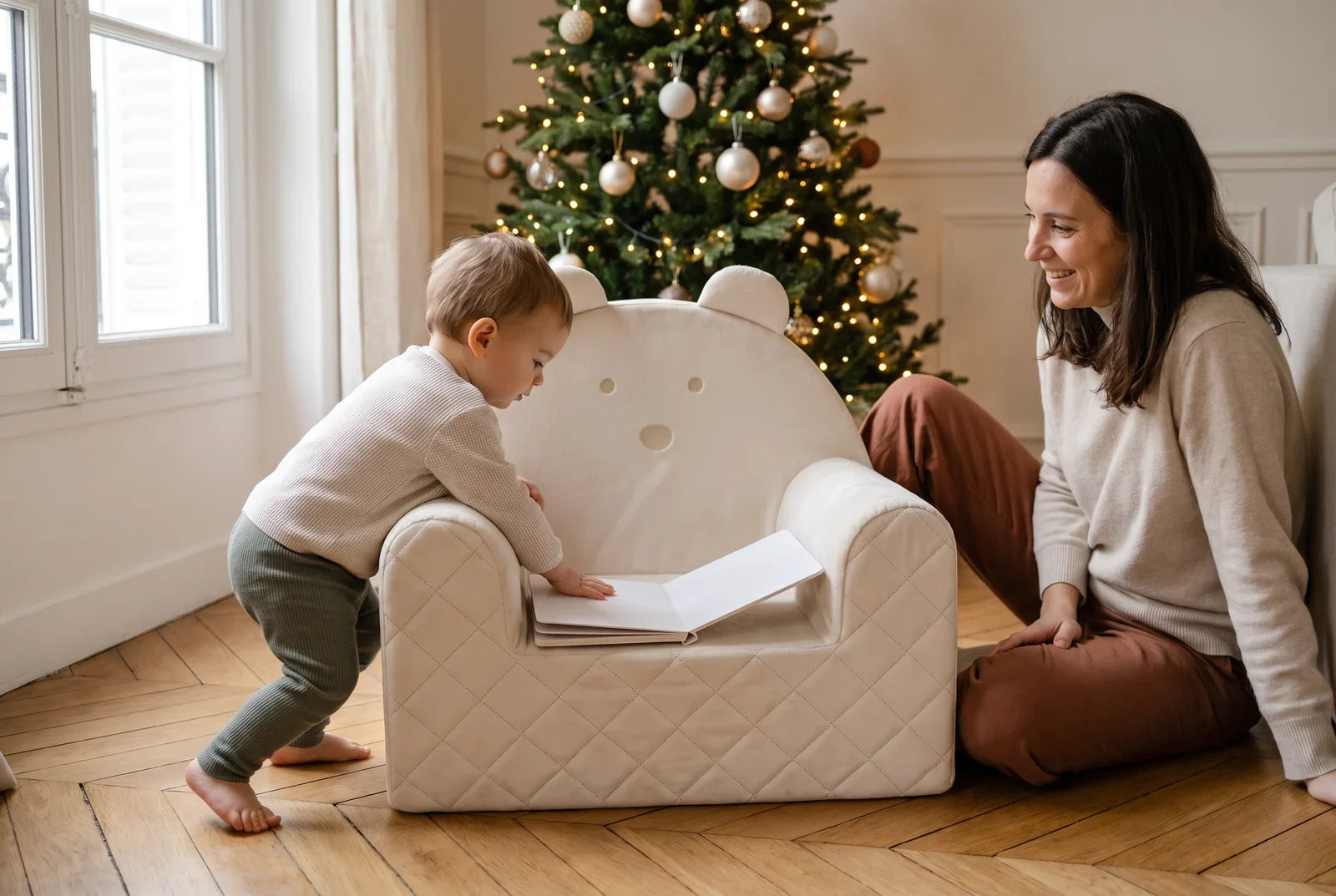 Tout-petit penché sur un livre posé sur un fauteuil bébé ourson en velours beige, un adulte assis à côté, près du sapin