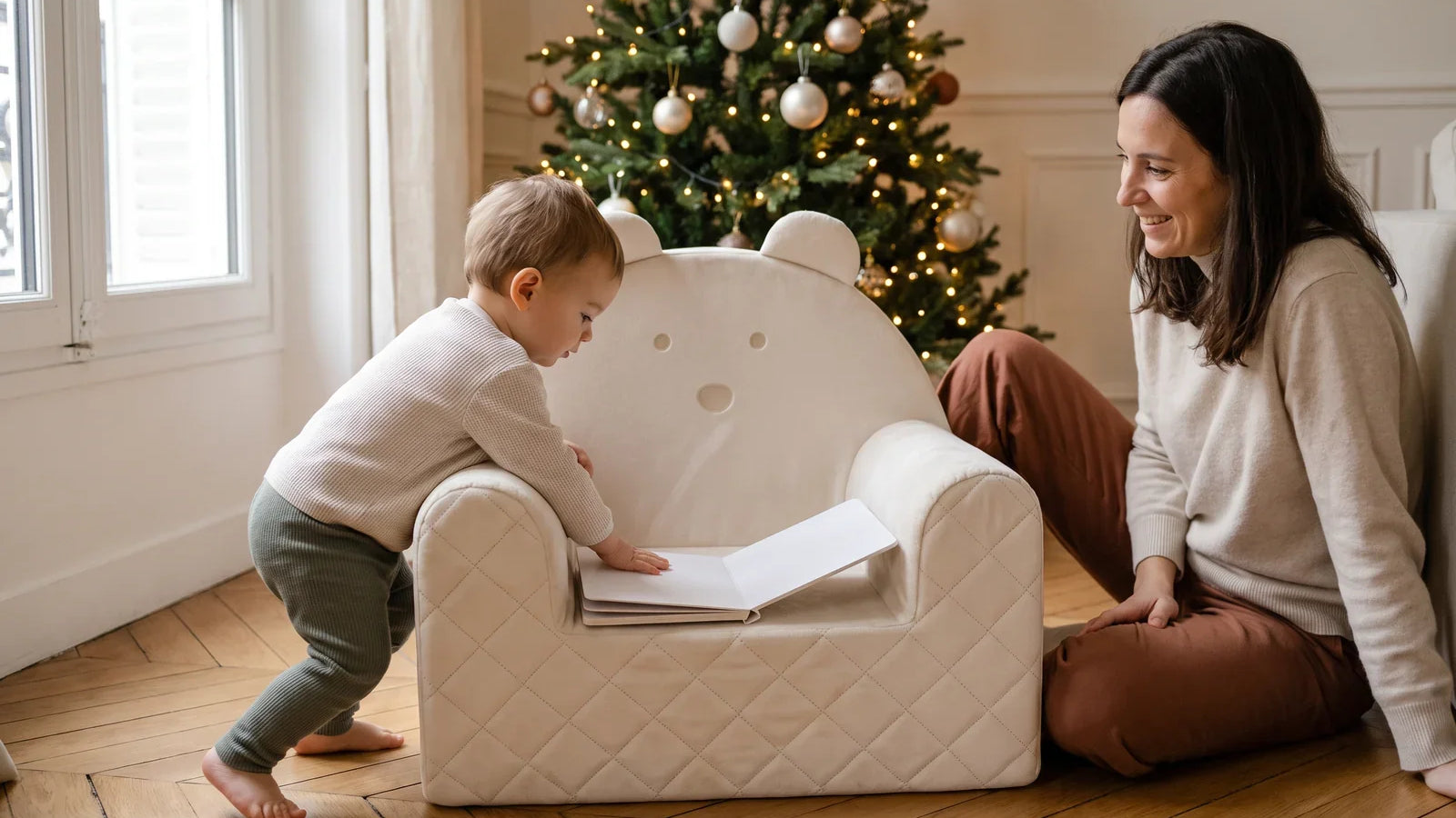 Tout-petit penché sur un livre posé sur un fauteuil bébé ourson en velours beige, un adulte assis à côté, près du sapin