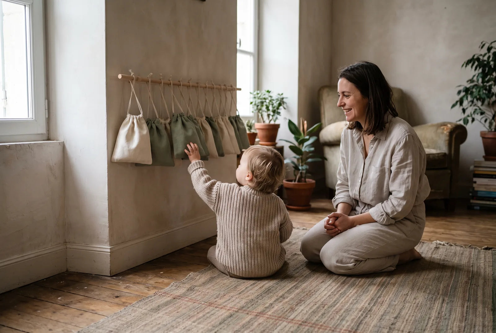 Enfant tendant la main vers des pochettes en tissu suspendues à une tringle, une femme souriante assise au sol à côté de lui