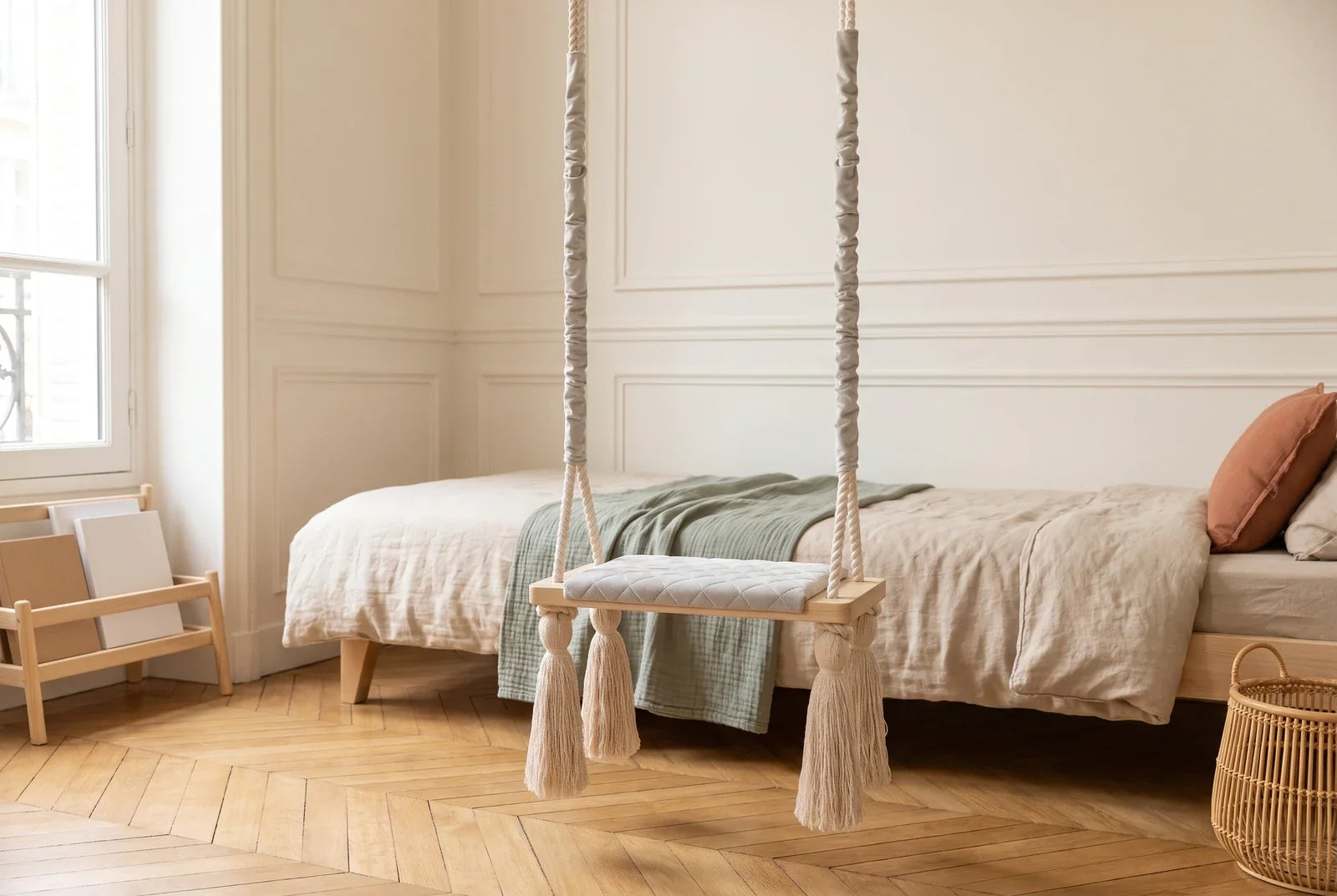 Balançoire planche en velours gris clair suspendue dans une chambre d’enfant lumineuse, devant un lit en bois