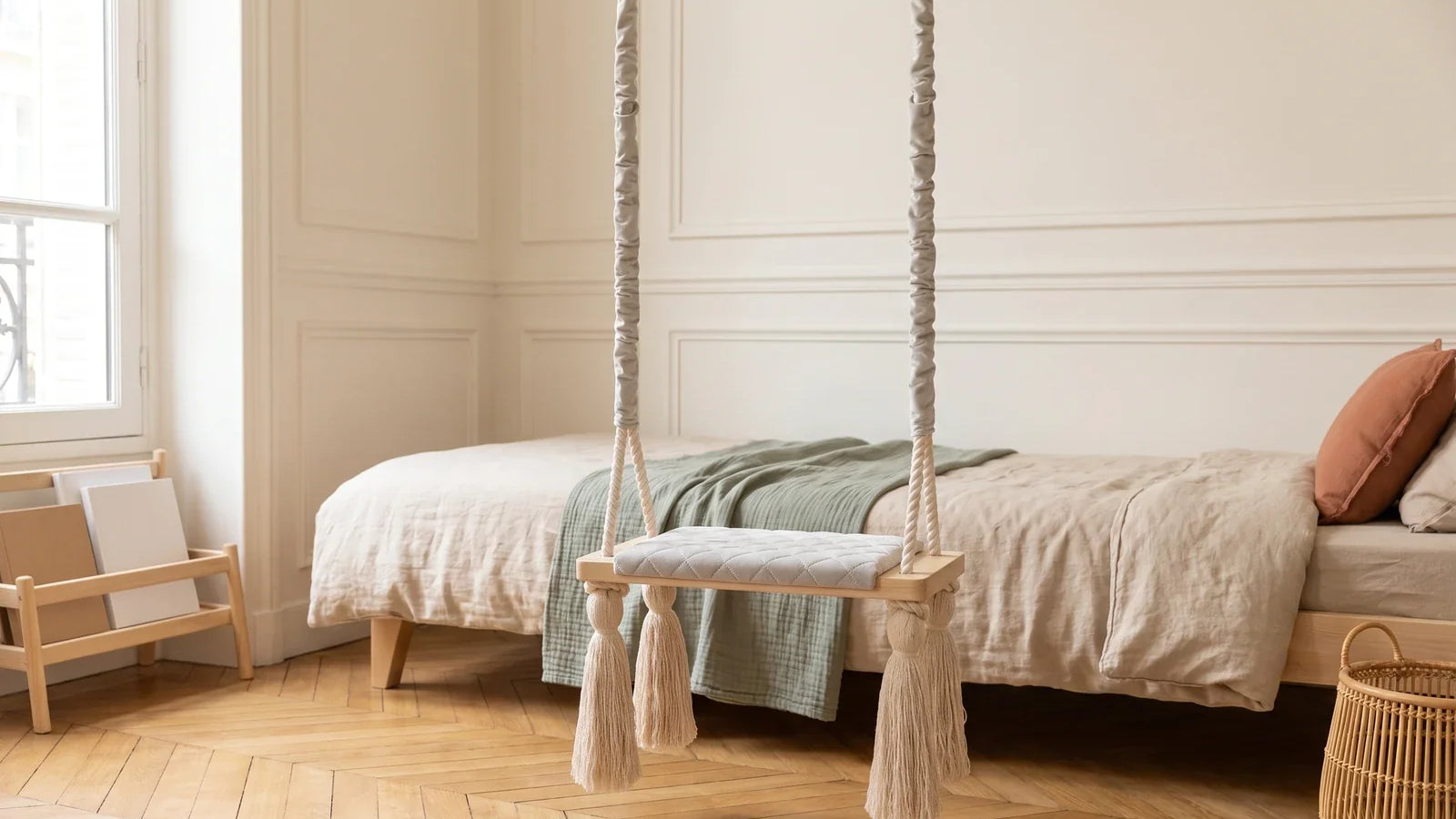 Balançoire planche en velours gris clair suspendue dans une chambre d’enfant lumineuse, devant un lit en bois