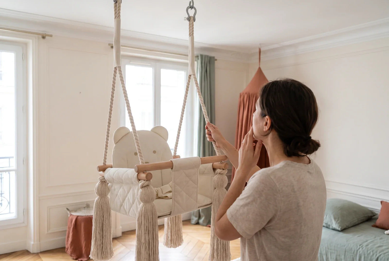 Une femme tire doucement sur la corde d’une balançoire bébé beige suspendue au plafond et lève les yeux vers le crochet