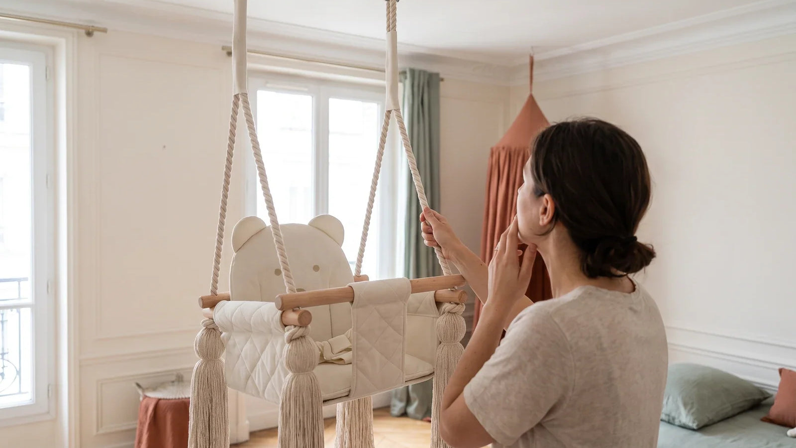 Une femme tire doucement sur la corde d’une balançoire bébé beige suspendue au plafond et lève les yeux vers le crochet