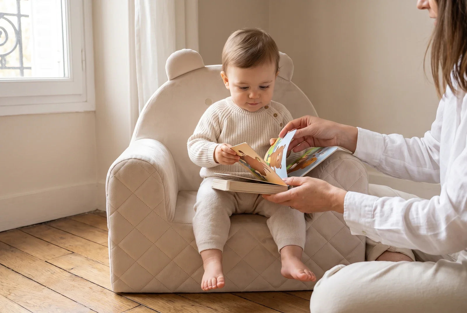 Enfant d'un an assis dans un fauteuil ourson beige en velours, avec un livre cartonné