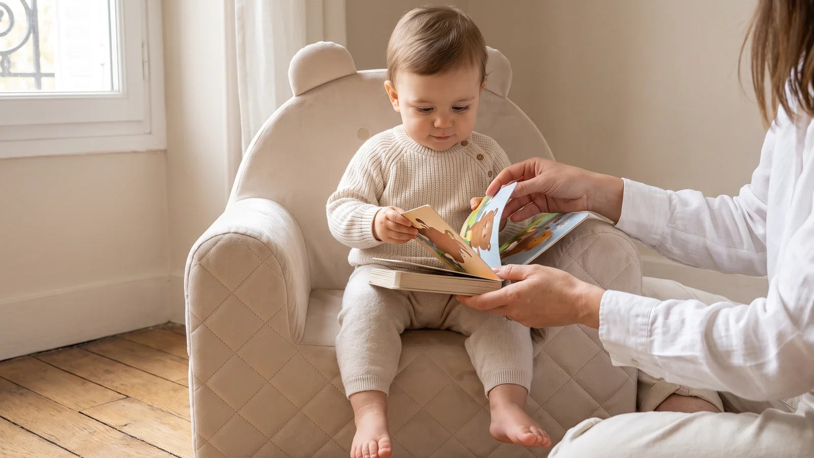 Enfant d'un an assis dans un fauteuil ourson beige en velours, avec un livre cartonné