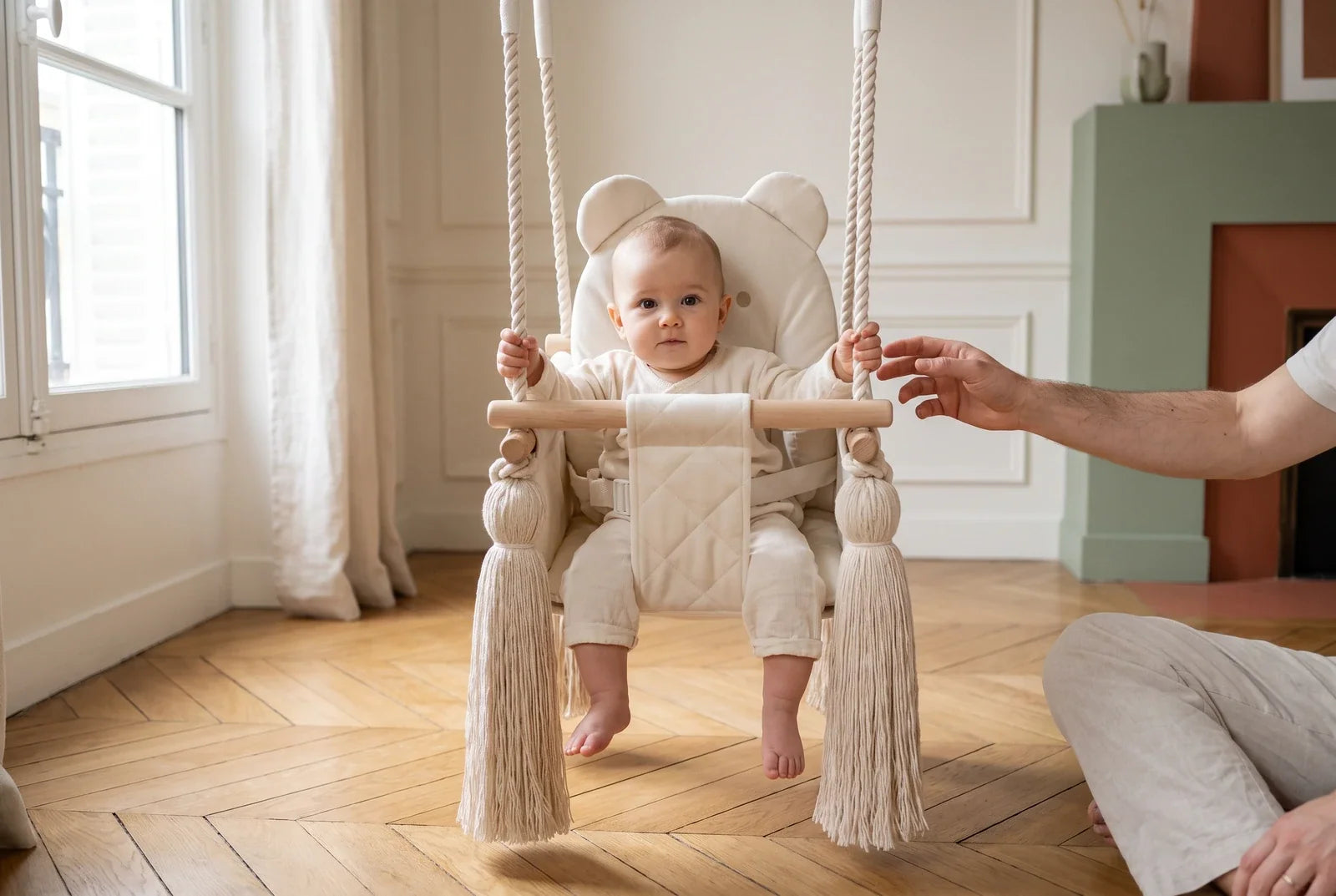 Un bébé d'environ 10 mois est assis dans la balançoire bébé en velours beige, un adulte assis au sol tend la main vers lui