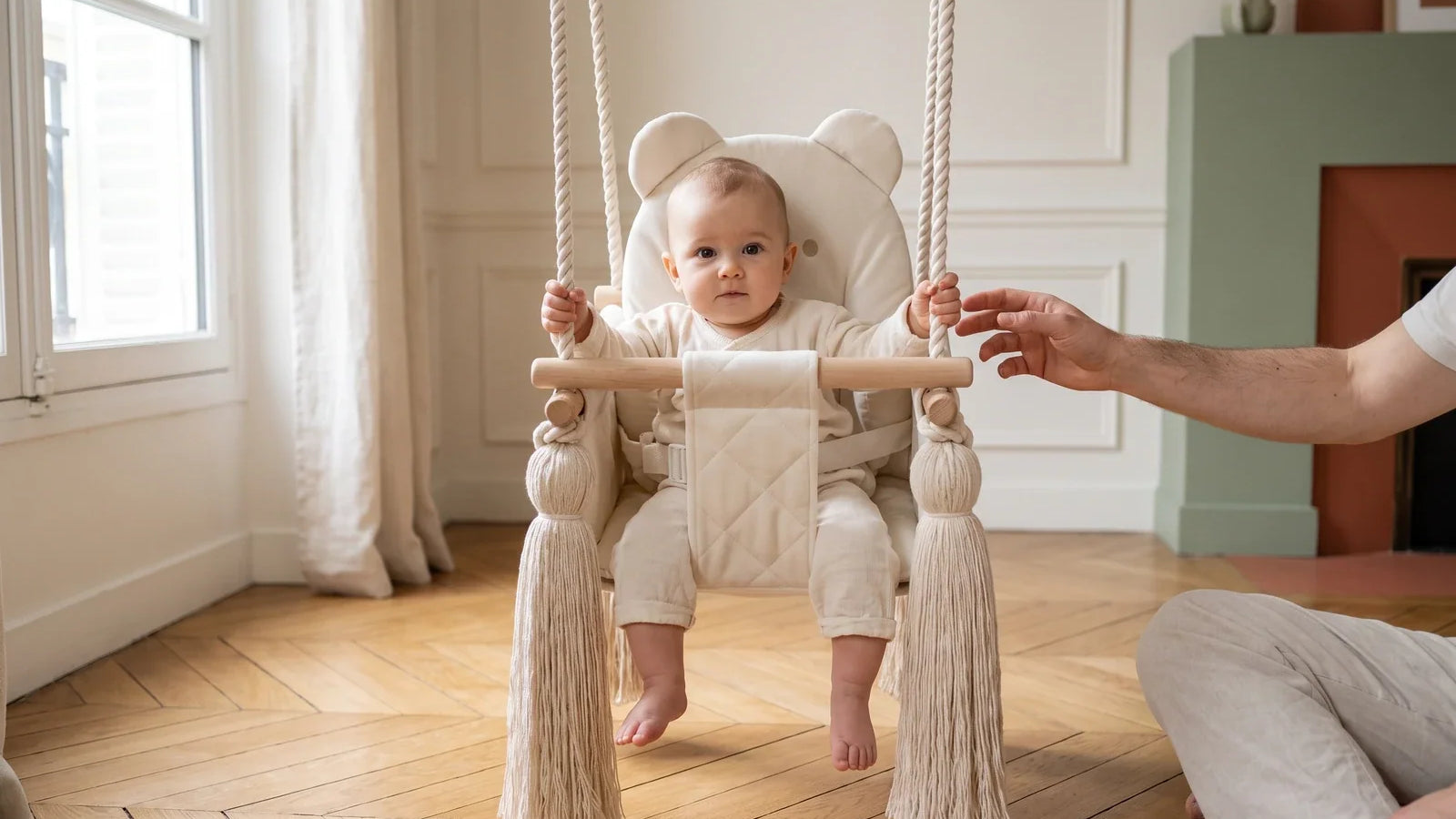Un bébé d'environ 10 mois est assis dans la balançoire bébé en velours beige, un adulte assis au sol tend la main vers lui