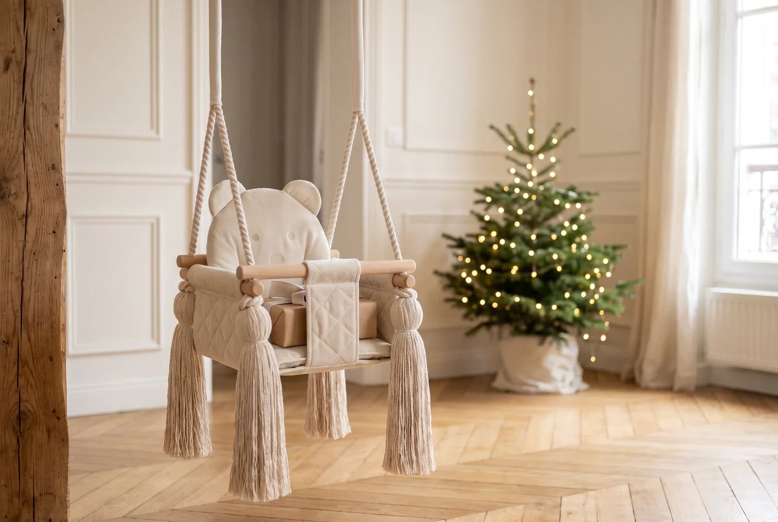 Balançoire bébé en velours beige avec un petit cadeau sur l’assise, devant un sapin de Noël dans un salon lumineux