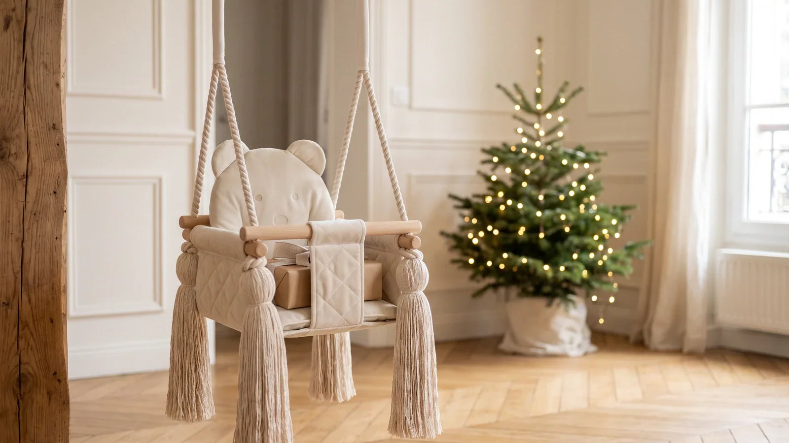 Balançoire bébé en velours beige avec un petit cadeau sur l’assise, devant un sapin de Noël dans un salon lumineux